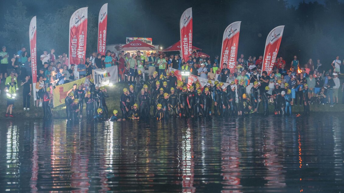Eine Gruppe von Menschen steht am Ufer eines Gewässers und posiert für ein Foto. Im Hintergrund sind festliche Banner und Lichter zu sehen. | © autxtri