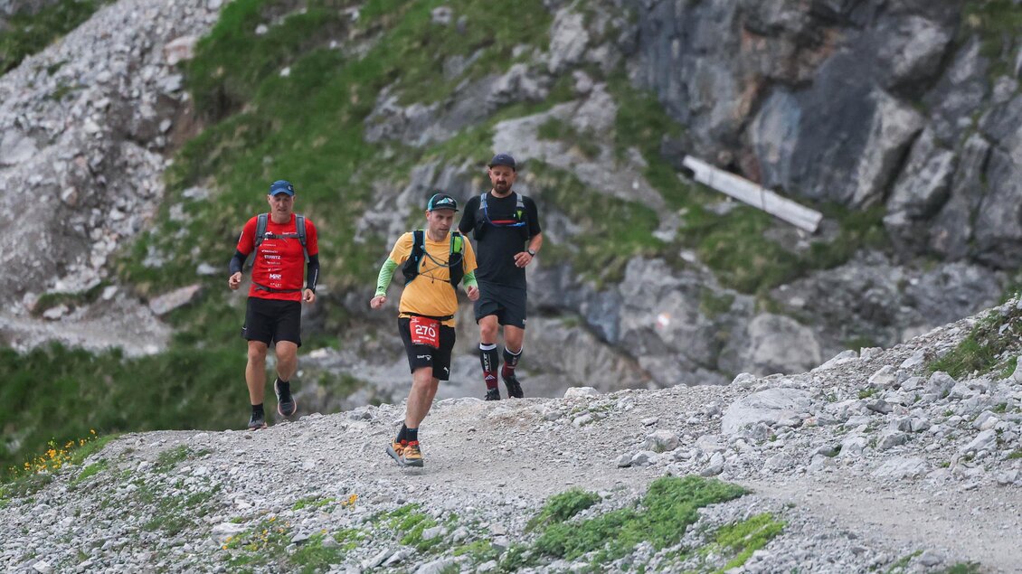 Drei Personen joggen auf einem schmalen Wanderweg in den Bergen. Die Umgebung ist felsig und grün, mit beeindruckenden Felsformationen im Hintergrund. | © autxtri