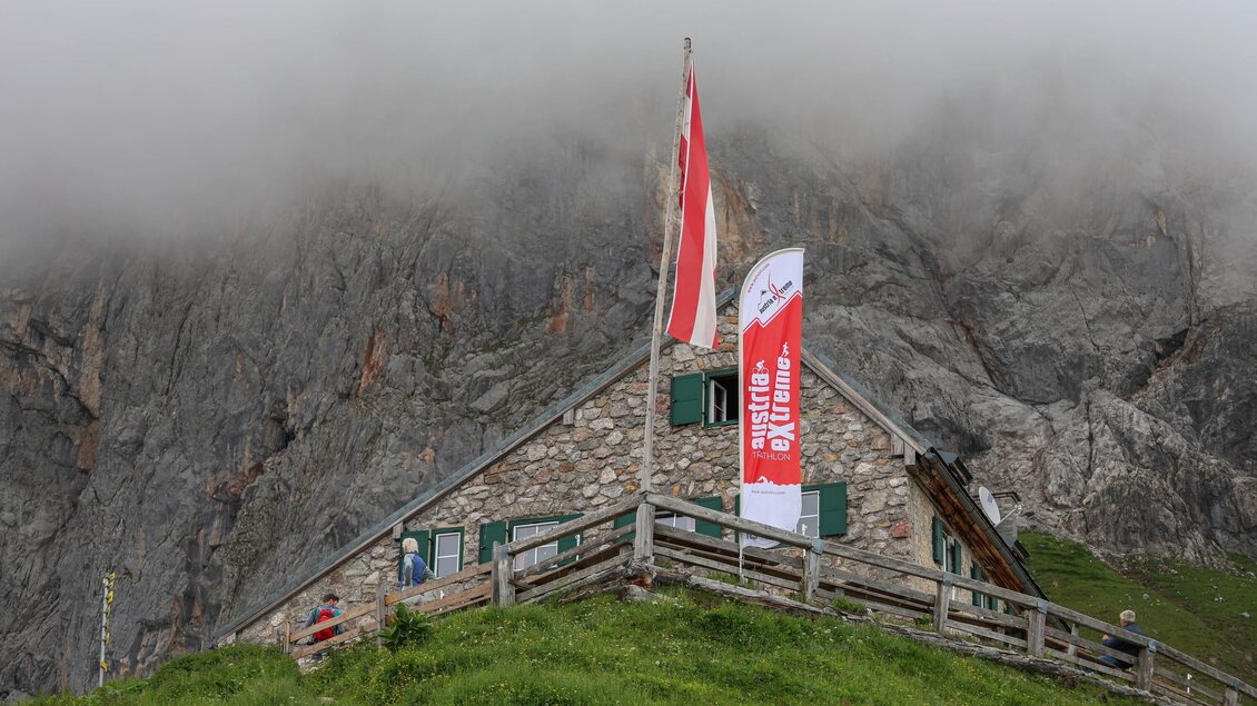 Eine Almhütte mit roten und weißen Fahnen in einer bewölkten Berglandschaft. Im Hintergrund sind steile Felsen sichtbar. | © autxtri