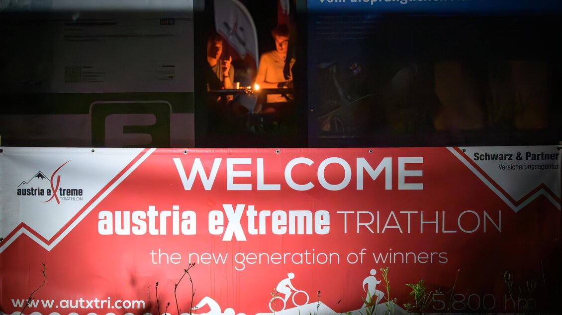 Banner der Abschlussveranstaltung des eXtreme Triathlons bei Nacht mit Teilnehmern im Hintergrund. | © Region Graz-Gerald Hirl