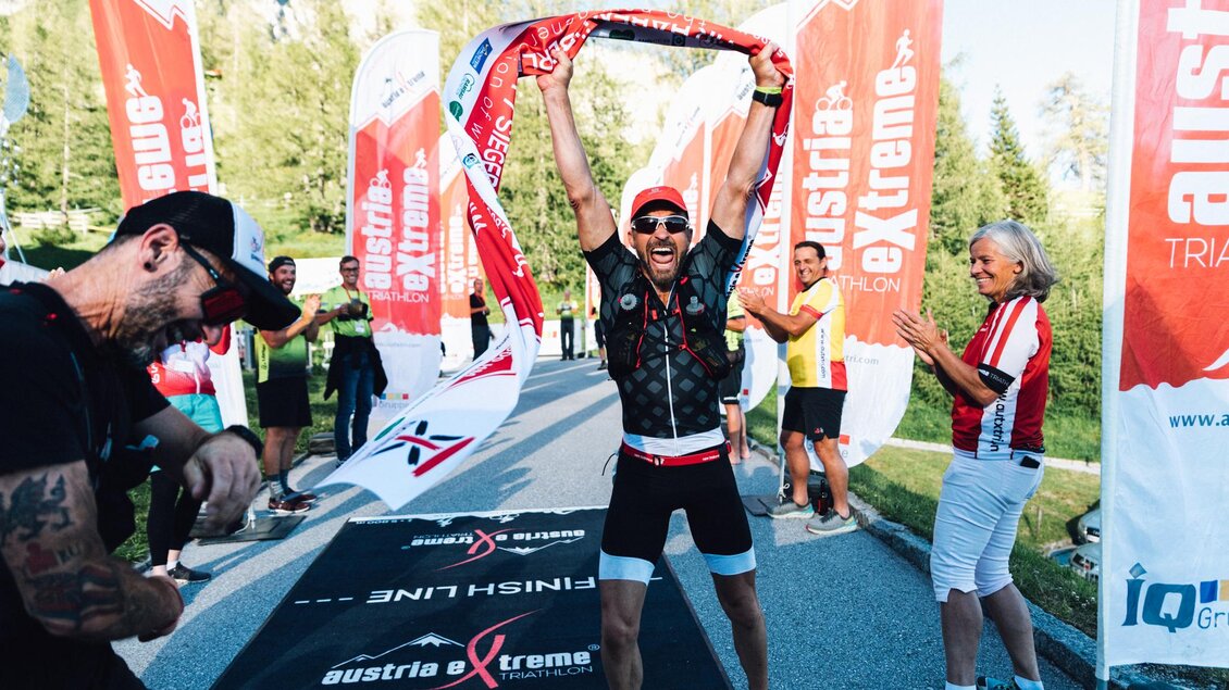 Ein begeisterter Sportler feiert den Zieleinlauf mit einer Flagge in der Hand. Im Hintergrund jubeln Zuschauer und weitere Teilnehmer. | © Austria eXtreme
