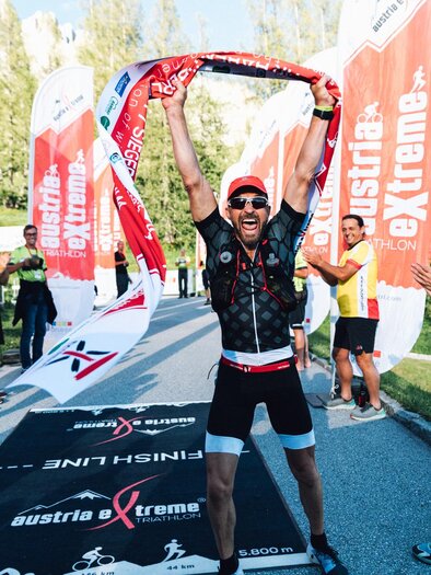 Ein begeisterter Sportler feiert den Zieleinlauf mit einer Flagge in der Hand. Im Hintergrund jubeln Zuschauer und weitere Teilnehmer. | © Austria eXtreme