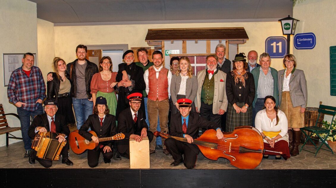 Eine Gruppe von Menschen posiert auf einer Bühnenkulisse. Einige tragen traditionelle Kleidung und halten Musikinstrumente. | © Erich Kainzinger