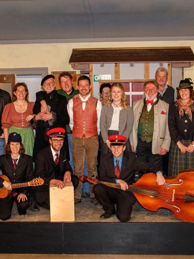 Eine Gruppe von Menschen posiert auf einer Bühnenkulisse. Einige tragen traditionelle Kleidung und halten Musikinstrumente. | © Erich Kainzinger