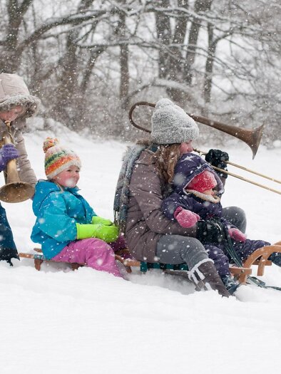 Eine Gruppe von Kindern und Erwachsenen spielt im Schnee. Einige fahren Schlitten, während andere Musikinstrumente spielen. | © (c)STVLW_Unterrainer