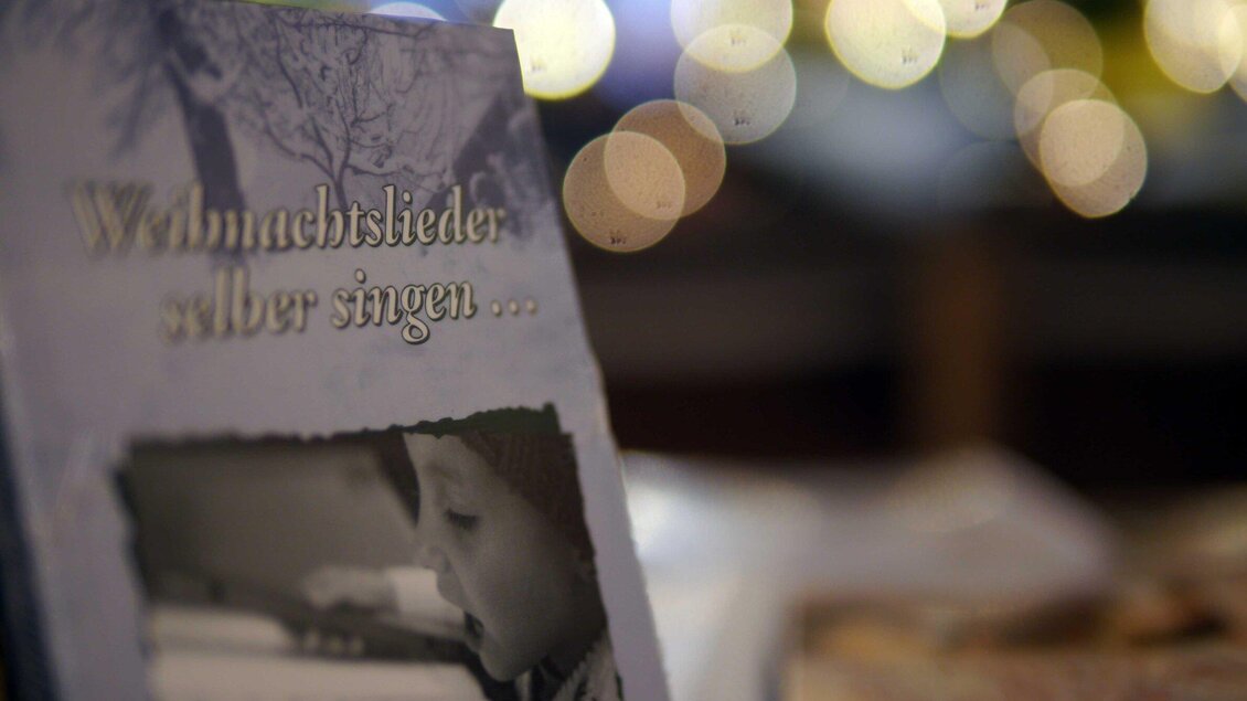Ein Buch über Weihnachtslieder steht auf einem Tisch. Die verschwommenen Lichter im Hintergrund erzeugen eine festliche Stimmung. | © Steirisches Volksliedwerk