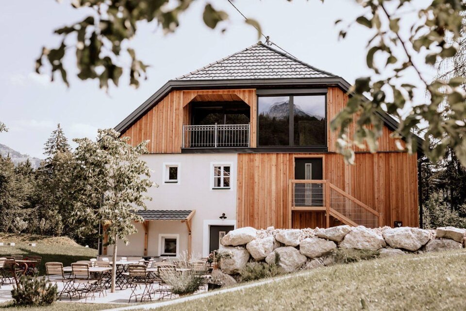 Eine Ansicht vom Eingang des Schutzhaus am Hohen Camper in Bad Aussee im steirischen Salzkammergut, die Fassade ist ein Mix aus traditionell und modern und mit viel Holz und Glas im Obergeschoss gestaltet, das Dach ist für die Region typisch geformt und grau gedeckt. Im Vordergrund sieht man eine nette Außenanlage mit Tischen und Stühlen unter einem kleinen Baum. | © SchutzhausAmHohenCamper