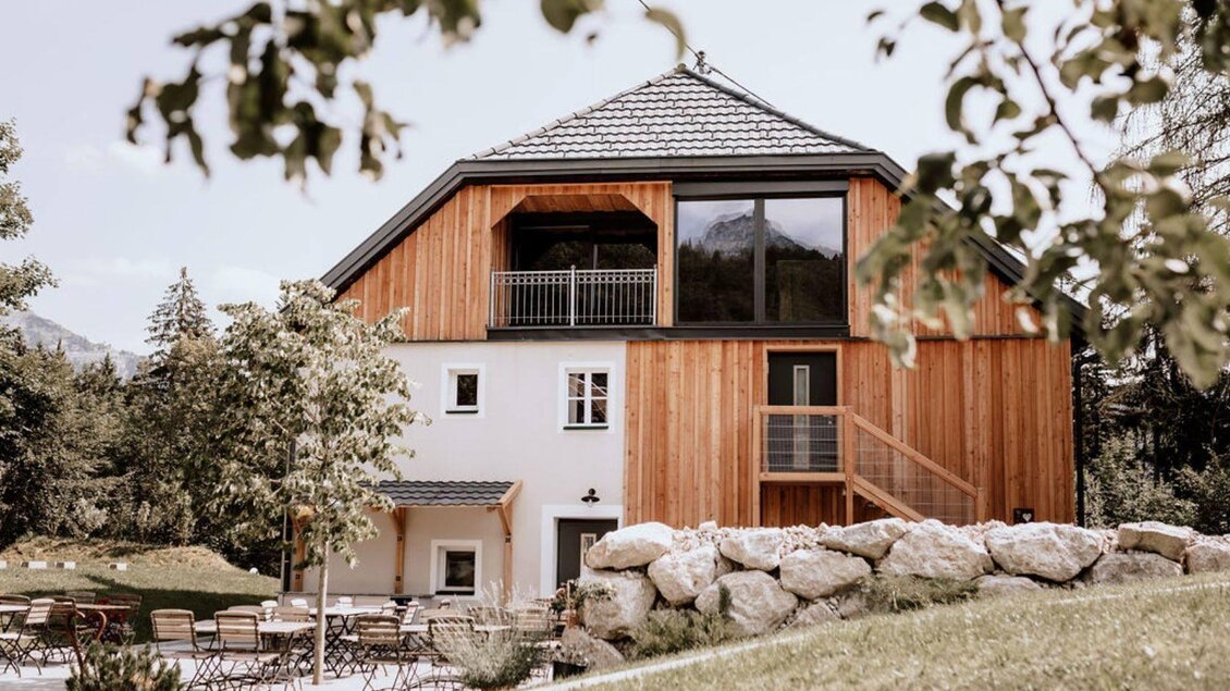 Eine Ansicht vom Eingang des Schutzhaus am Hohen Camper in Bad Aussee im steirischen Salzkammergut, die Fassade ist ein Mix aus traditionell und modern und mit viel Holz und Glas im Obergeschoss gestaltet, das Dach ist für die Region typisch geformt und grau gedeckt. Im Vordergrund sieht man eine nette Außenanlage mit Tischen und Stühlen unter einem kleinen Baum. | © SchutzhausAmHohenCamper