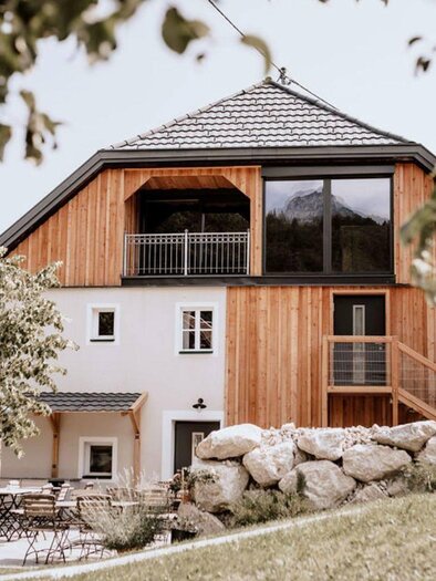 A view from the entrance of the "Schutzhaus am Hohen Camper" in Bad Aussee in the Styrian Salzkammergut, the façade is a mix of traditional and modern design, with plenty of wood and glass on the upper floor, the roof is typically shaped for the region and covered in gray. In the foreground, you can see a nice outdoor area with tables and chairs under a small tree. | © SchutzhausAmHohenCamper