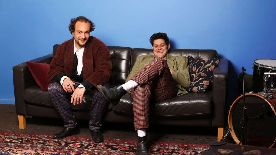Zwei Männer sitzen entspannt auf einer Couch vor einer blauen Wand. Der eine trägt einen Mantel und der andere ein sportliches Outfit mit karierten Hosen. | © das G'MISCH