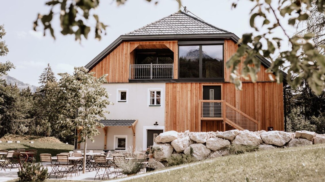 Das moderne Schutzhaus am hohen Camper mit Holzfassade und großen Fenstern befindet sich mittig vom Bild. Im Vordergrund sind Gartenmöbel und Grünflächen zu sehen.
 | © das Schutzhaus am hohen Camper, Bad Aussee