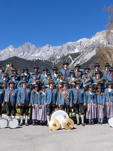 Eine große Gruppe von Menschen in traditioneller Kleidung steht im Freien vor einer beeindruckenden Berglandschaft. Sie halten Musikinstrumente und lächeln in die Kamera. | © Harald Steiner