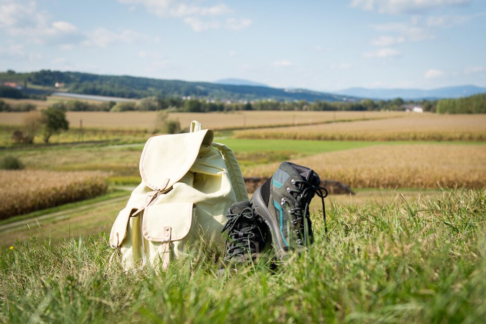 Ein Rucksack und ein Paar Wanderschuhe stehen auf einer Wiese. Im Hintergrund ist ein weitläufiges Feld und ein blauer Himmel zu sehen. | © Kurkommission Bad Blumau