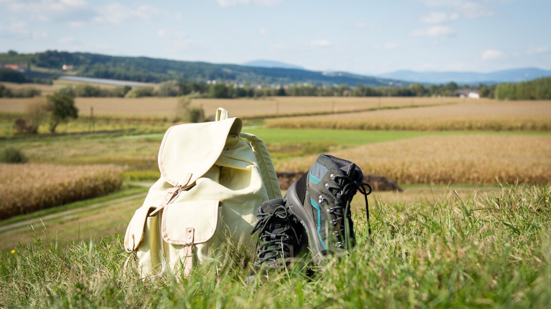 Ein Rucksack und ein Paar Wanderschuhe stehen auf einer Wiese. Im Hintergrund ist ein weitläufiges Feld und ein blauer Himmel zu sehen. | © Kurkommission Bad Blumau