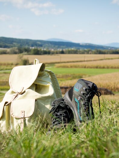 A backpack and a pair of hiking shoes are standing on a meadow. In the background, a vast field and a blue sky can be seen. | © Kurkommission Bad Blumau