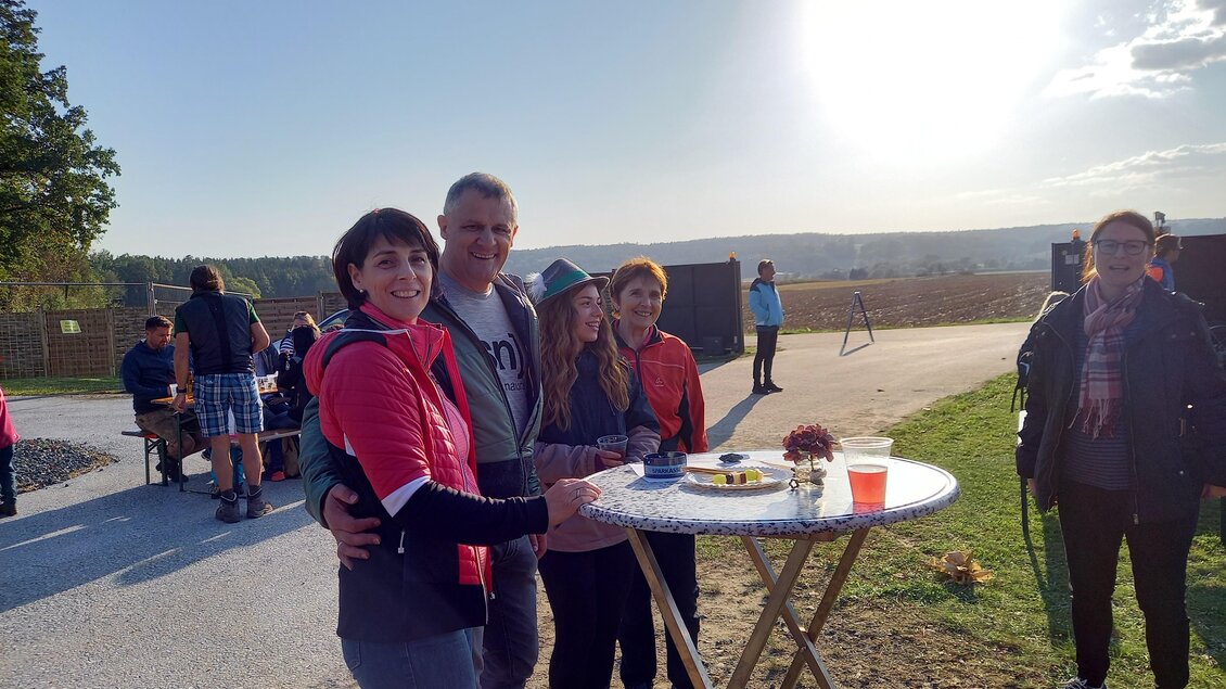 Eine Gruppe von vier Personen steht an einem Tisch im Freien. Im Hintergrund sind eine malerische Landschaft und eine Sonnenschein-Szene zu sehen.