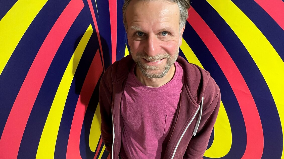 Ein Mann steht vor einem bunten, psychedelischen Hintergrund mit Streifen in Gelb, Blau und Rot. Er lächelt und trägt ein lässiges Outfit. | © © Marva Held