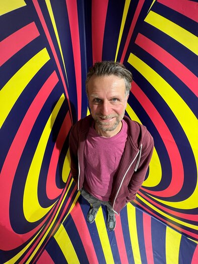 Ein Mann steht vor einem bunten, psychedelischen Hintergrund mit Streifen in Gelb, Blau und Rot. Er lächelt und trägt ein lässiges Outfit. | © © Marva Held