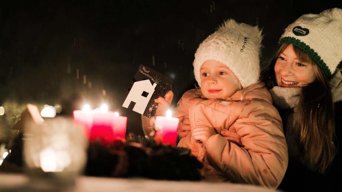 Eine Mutter sitzt mit ihrem Kind an einem Tisch, der mit Kerzen geschmückt ist. Beide tragen warme Mützen und genießen die winterliche Atmosphäre. | © Lupi Spuma