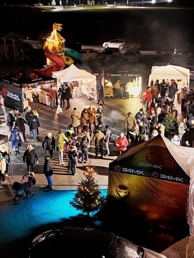 **Atmospheric Advent Market in Bad Waltersdorf:**
From a bird's-eye view, a festively lit Advent market reveals itself, buzzing with numerous visitors. Decorated Christmas trees, market stalls, and warm lights create a cozy atmosphere, inviting people to enjoy, chat, and linger together. | © Faiss Rahmen