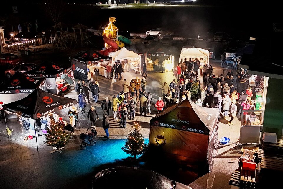 **Stimmungsvoller Adventmarkt in Bad Waltersdorf:**
Aus der Vogelperspektive zeigt sich ein festlich beleuchteter Adventmarkt mit zahlreichen Besucherinnen und Besuchern. Geschmückte Weihnachtsbäume, Marktstände und warme Lichter sorgen für eine gemütliche Atmosphäre und laden zum gemeinsamen Genießen, Plaudern und Verweilen ein. | © Faiss Rahmen