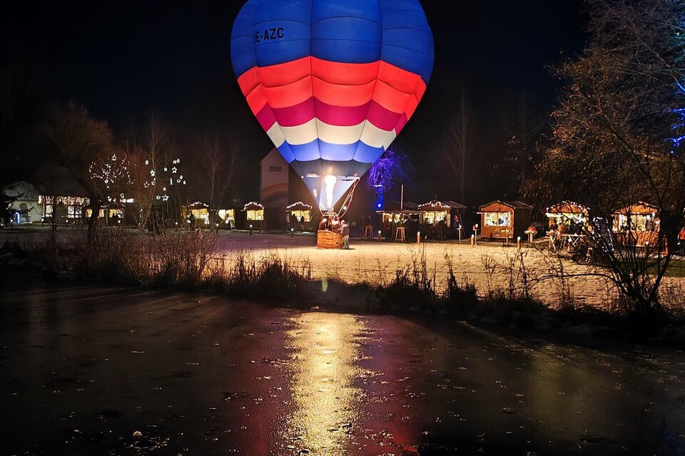 Ein bunter Heißluftballon schwebt über einem gefrorenen Teich in der Nacht. Um den Teich herum sind festlich aufgerichtete Stände zu sehen. | © ©Lederer Josef/Thermen-&Vulkanland