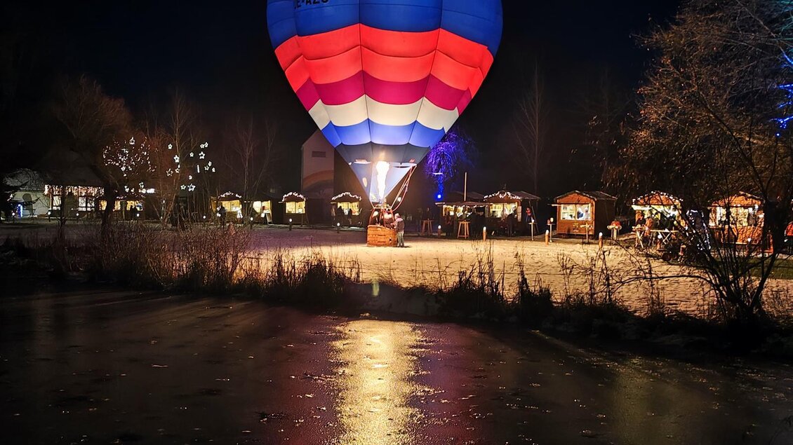 Ein bunter Heißluftballon schwebt über einem gefrorenen Teich in der Nacht. Um den Teich herum sind festlich aufgerichtete Stände zu sehen. | © ©Lederer Josef/Thermen-&Vulkanland