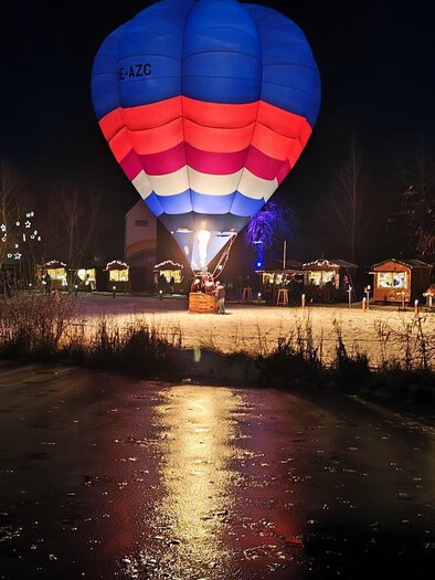 Ein bunter Heißluftballon schwebt über einem gefrorenen Teich in der Nacht. Um den Teich herum sind festlich aufgerichtete Stände zu sehen. | © ©Lederer Josef/Thermen-&Vulkanland