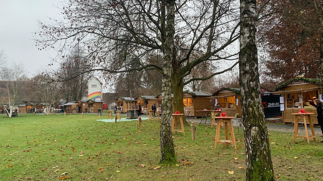 Eine Grünfläche mit mehreren Holzhütten und Tischen unter Bäumen. Der Himmel ist bewölkt und die Umgebung hat herbstliche Farben.