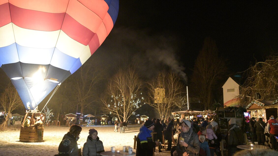 Ein bunter Heißluftballon leuchtet in der Nacht. Menschen versammeln sich um Stände und genießen die festliche Atmosphäre. | © Lederer