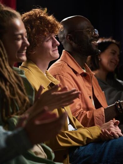 Ein Publikum sitzt im Saal und schaut auf die Bühne. Bunte Lichter und eine musikalische Darbietung sorgen für eine lebhafte Stimmung. | © AdobeStock