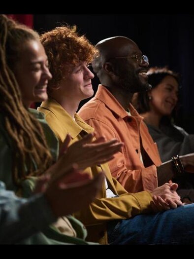 An audience is sitting in the hall and looking at the stage. Colorful lights and a musical performance create a lively atmosphere. | © AdobeStock