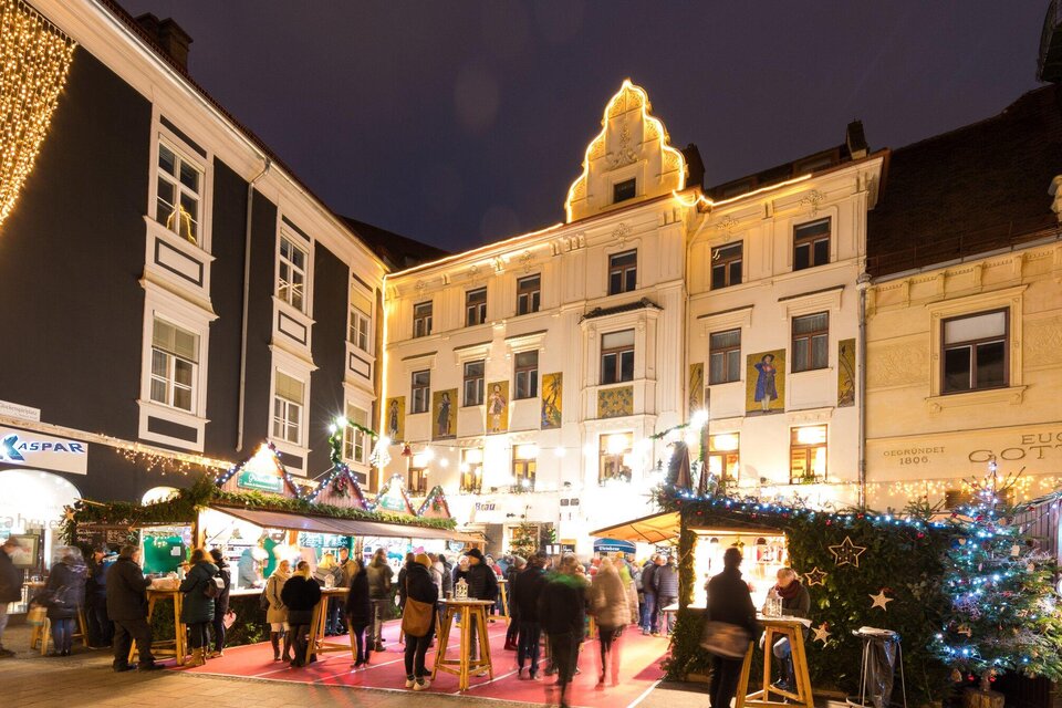 Ein festlicher Weihnachtsmarkt mit bunten Ständen und fröhlichen Menschen. Die Gebäude sind schön beleuchtet und schaffen eine gemütliche Atmosphäre. | © Graz Tourismus - Harry Schiffer