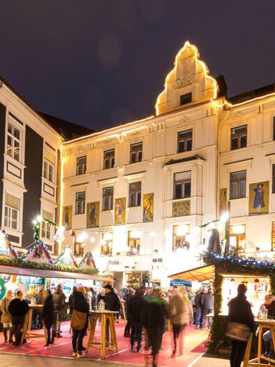 A festive Christmas market with colorful stalls and cheerful people. The buildings are beautifully lit and create a cozy atmosphere. | © Graz Tourismus - Harry Schiffer