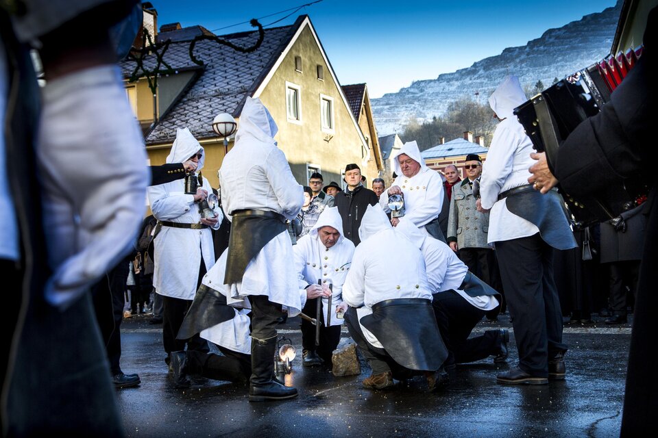 Eine Gruppe von Menschen in traditionellen Kostümen führt eine Zeremonie auf einer Straße durch. Im Hintergrund sind Häuser und eine Berglandschaft sichtbar. | © August Zoebl