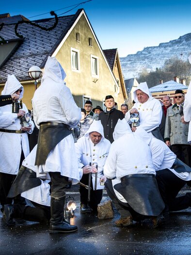 A group of people in traditional costumes is performing a ceremony on a street. In the background, houses and a mountain landscape are visible. | © August Zoebl