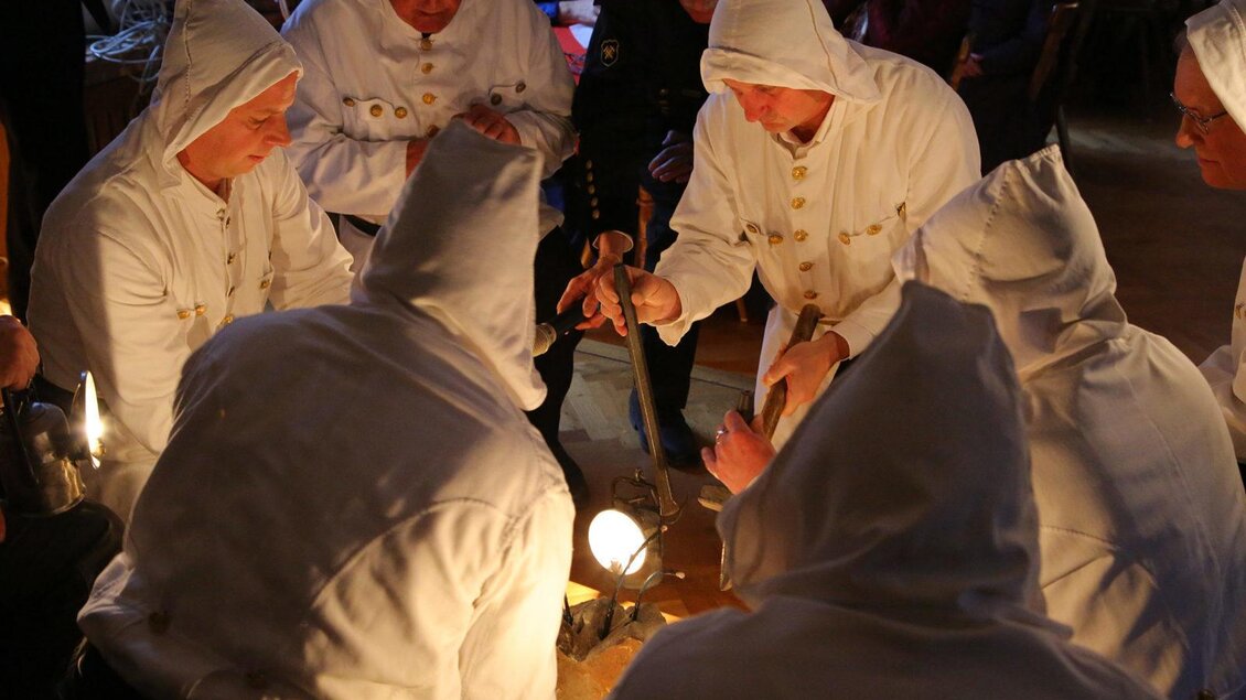 Eine Gruppe von Personen in weißen Kapuzenkleidern versammelt sich um einen Tisch. Sie scheinen an einer Zeremonie oder Ritual beteiligt zu sein, während sie ein Licht betrachten. | © Kulturquartier Leoben