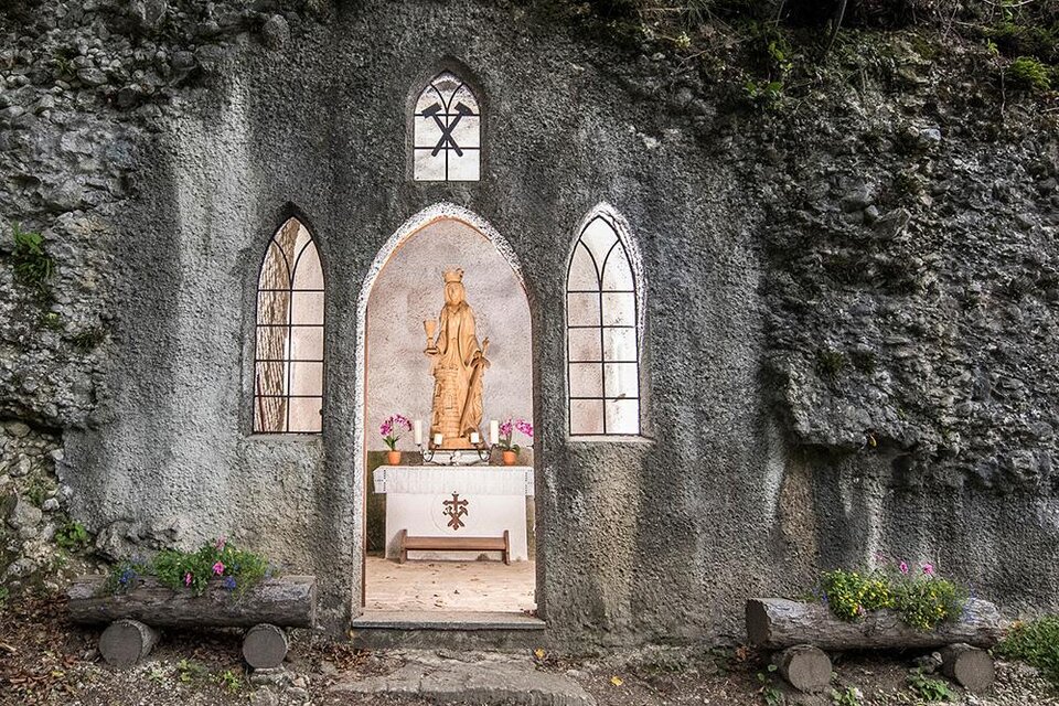 Eine kleine Kapelle in einer Felsenwand mit einer Holzstatue. Vor dem Altar stehen Blumen und Bänke aus Holz. | © Pressberger