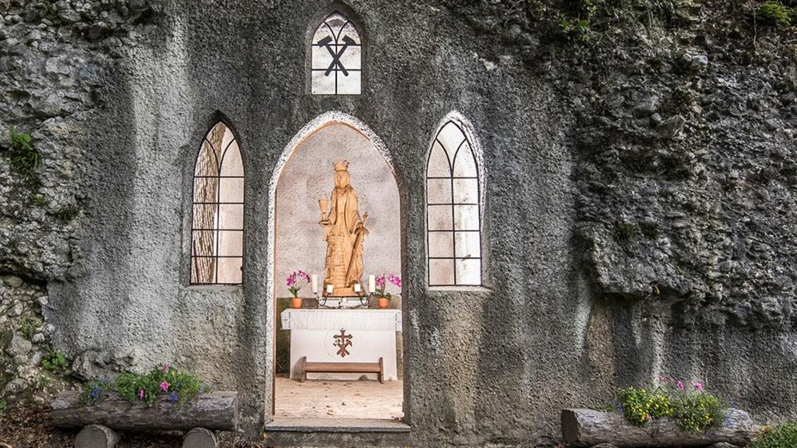 Eine kleine Kapelle in einer Felsenwand mit einer Holzstatue. Vor dem Altar stehen Blumen und Bänke aus Holz. | © Pressberger
