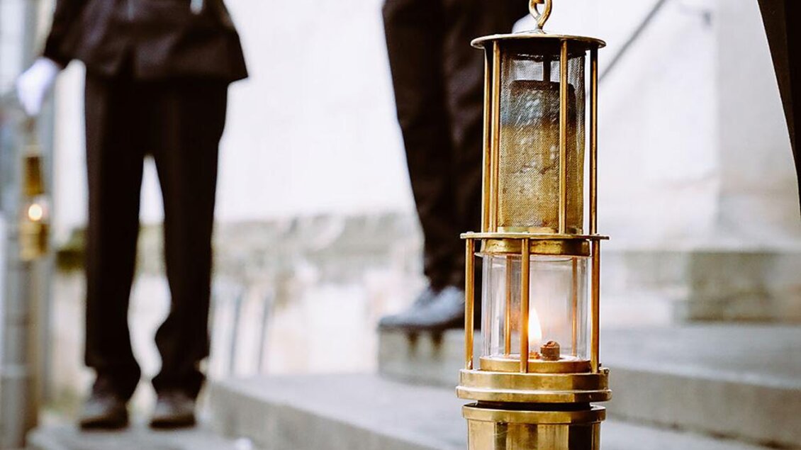 Eine antike Laterne aus Messing mit einer flackernden Flamme steht im Vordergrund. Im Hintergrund sind Männer in Uniformen zu sehen. | © Bild: Russold