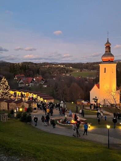 Menschen besuchen den Bartholomäer Adventmarkt mit Kirche und Lichtern in der Dämmerung. | © Josef Birnstingl