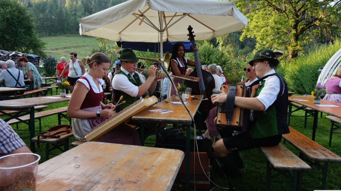 Eine musikalische Darbietung im Freien mit mehreren Musikern, die traditionelle Instrumente spielen. Im Hintergrund sind vorbeigehende Gäste und eine grüne Landschaft sichtbar.