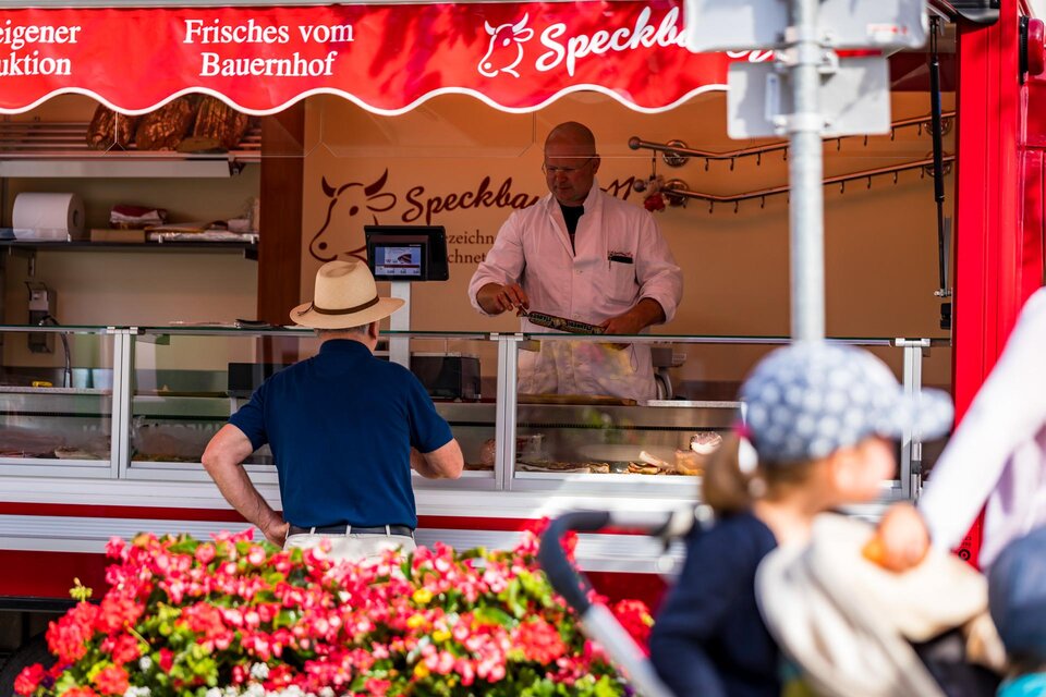 Ein Verkaufsstand mit einem Verkäufer, der frische Produkte anbietet. Im Vordergrund sieht man Blumen und einen Kunden, der interessiert zuschaut.