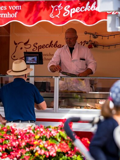 A sales booth with a seller offering fresh produce. In the foreground, flowers can be seen and a customer is watching with interest.