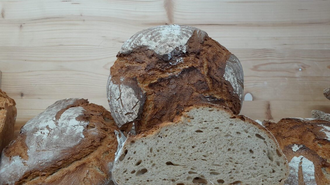 Ein rustikales Brot mit knuspriger Kruste und weichem Inneren. Es liegt auf einem Holzregal, umgeben von weiteren Broten. | © Bauernmarkt Riegersburg