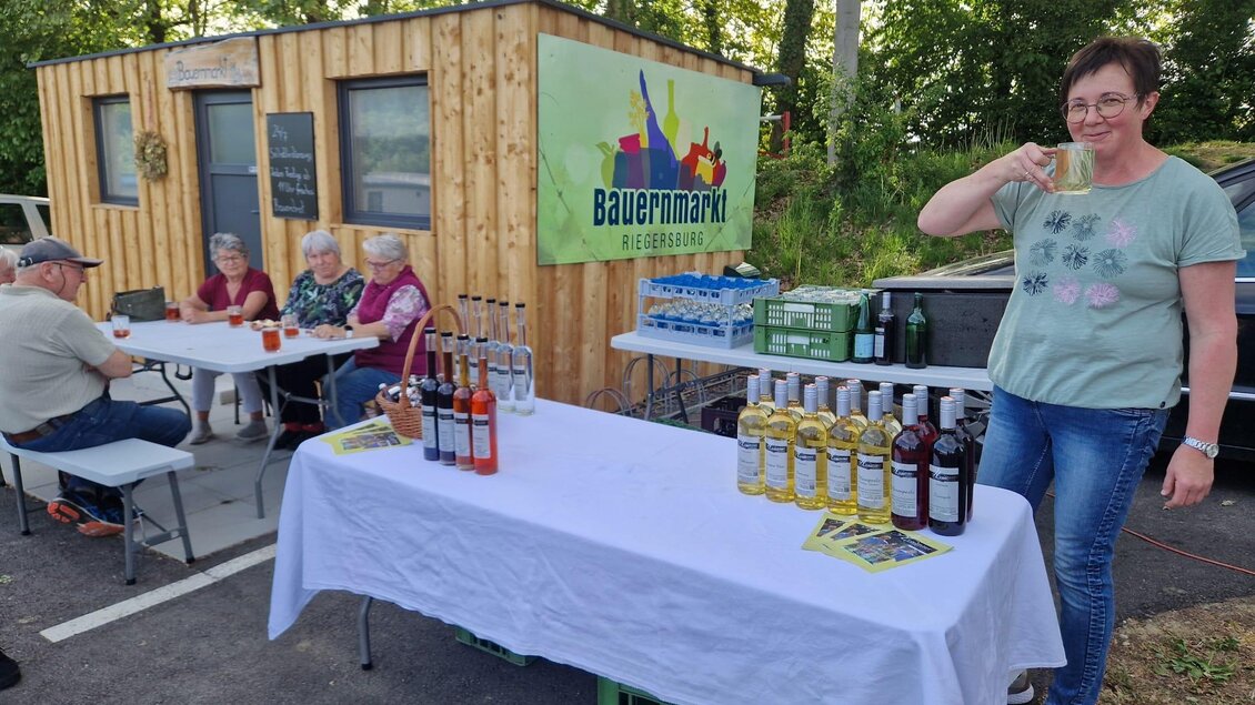 Auf einem Bauernmarkt stehen Tische mit Getränken und eine zufriedene Verkäuferin. Im Hintergrund sitzen einige Besucher und genießen die Atmosphäre. | © Camping Resort Riegersburg