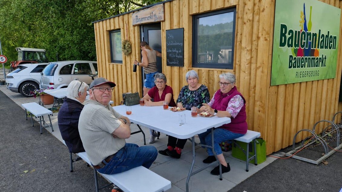 Eine Gruppe von Menschen sitzt an einem Tisch vor einem kleinen Bauernladen. Im Hintergrund sind einige Autos und Bäume zu sehen. | © Camping Resort Riegersburg
