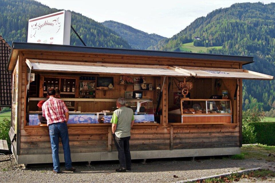 Ein Holzstand mit regionalen Produkten inmitten einer malerischen Landschaft. Zwei Männer betrachten die Angebote am Verkaufsstand. | © Region Murau/Famile Zeiner/