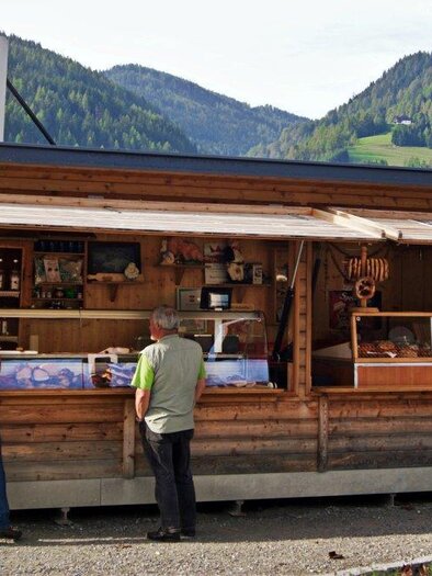Ein Holzstand mit regionalen Produkten inmitten einer malerischen Landschaft. Zwei Männer betrachten die Angebote am Verkaufsstand. | © Region Murau/Famile Zeiner/