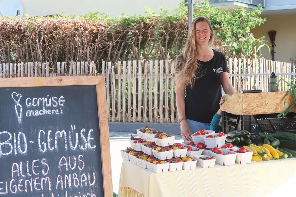 Eine Frau steht an einem Stand mit frischem bio Gemüse und Obst. Im Hintergrund ist eine Holzwand und Sonnenlicht zu sehen. | © Doll